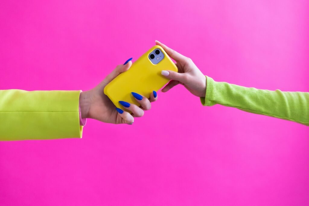 Colorful studio image of hands exchanging a yellow smartphone against a pink backdrop.